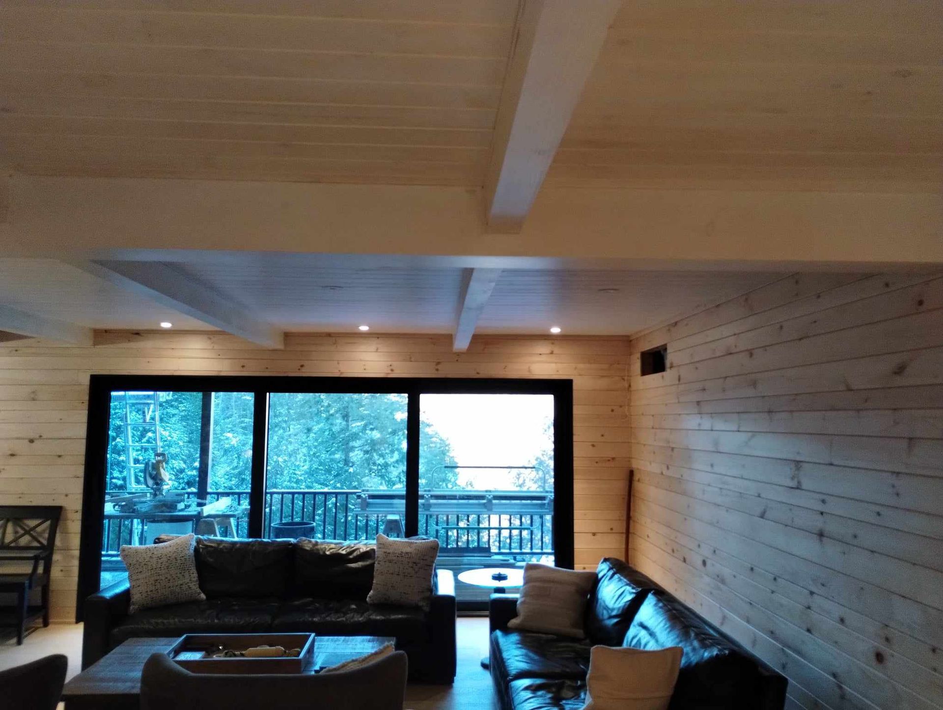 Salon moderne avec murs en bois, canapés foncés et une grande fenêtre donnant sur les arbres et une terrasse.
