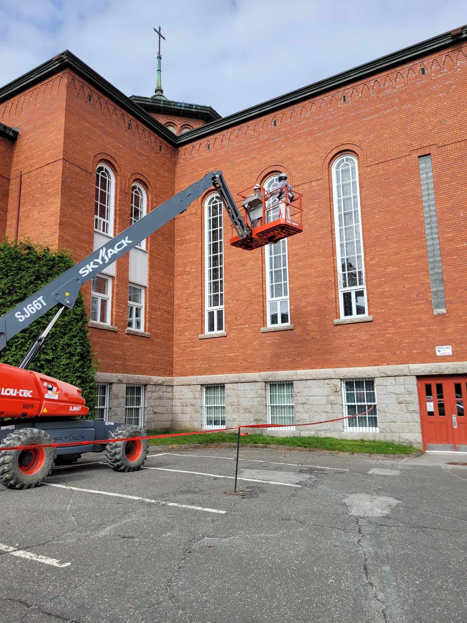 Ouvrier dans une nacelle rouge réparant un grand bâtiment en briques à côté d'un toit ressemblant à celui d'une église