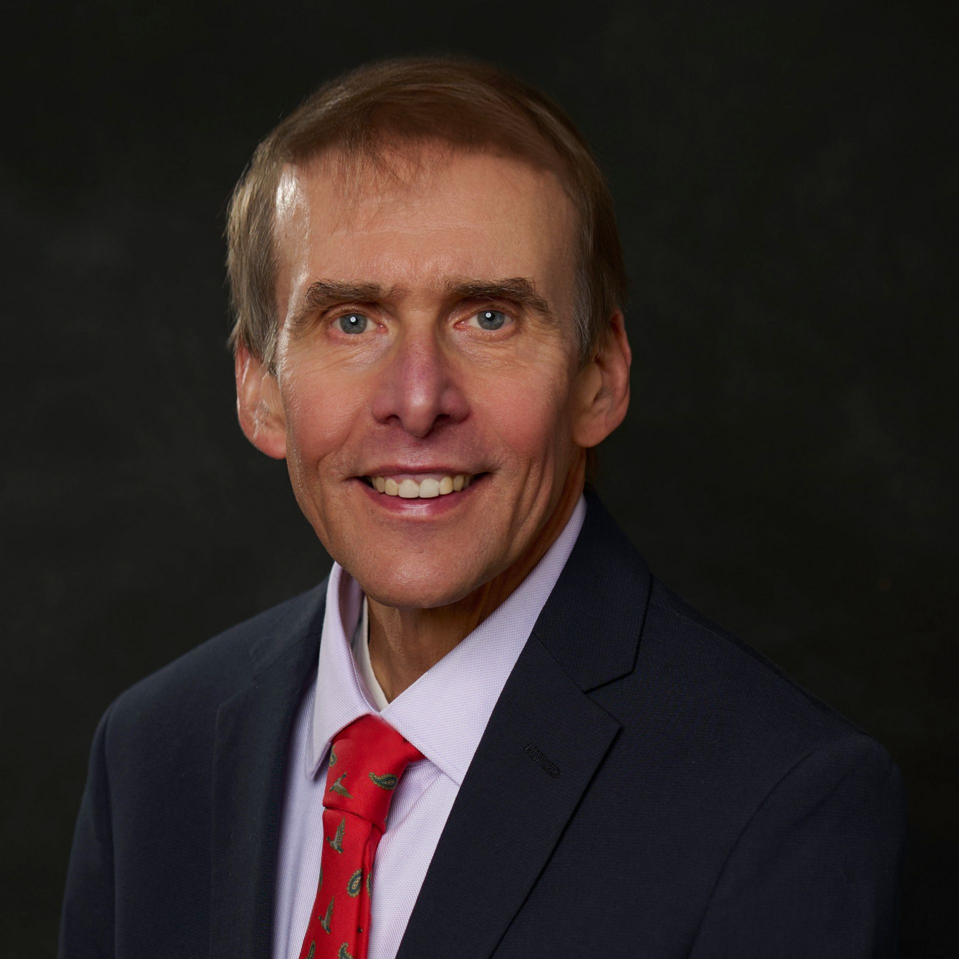 Man in suit and tie smiling, against a dark background.