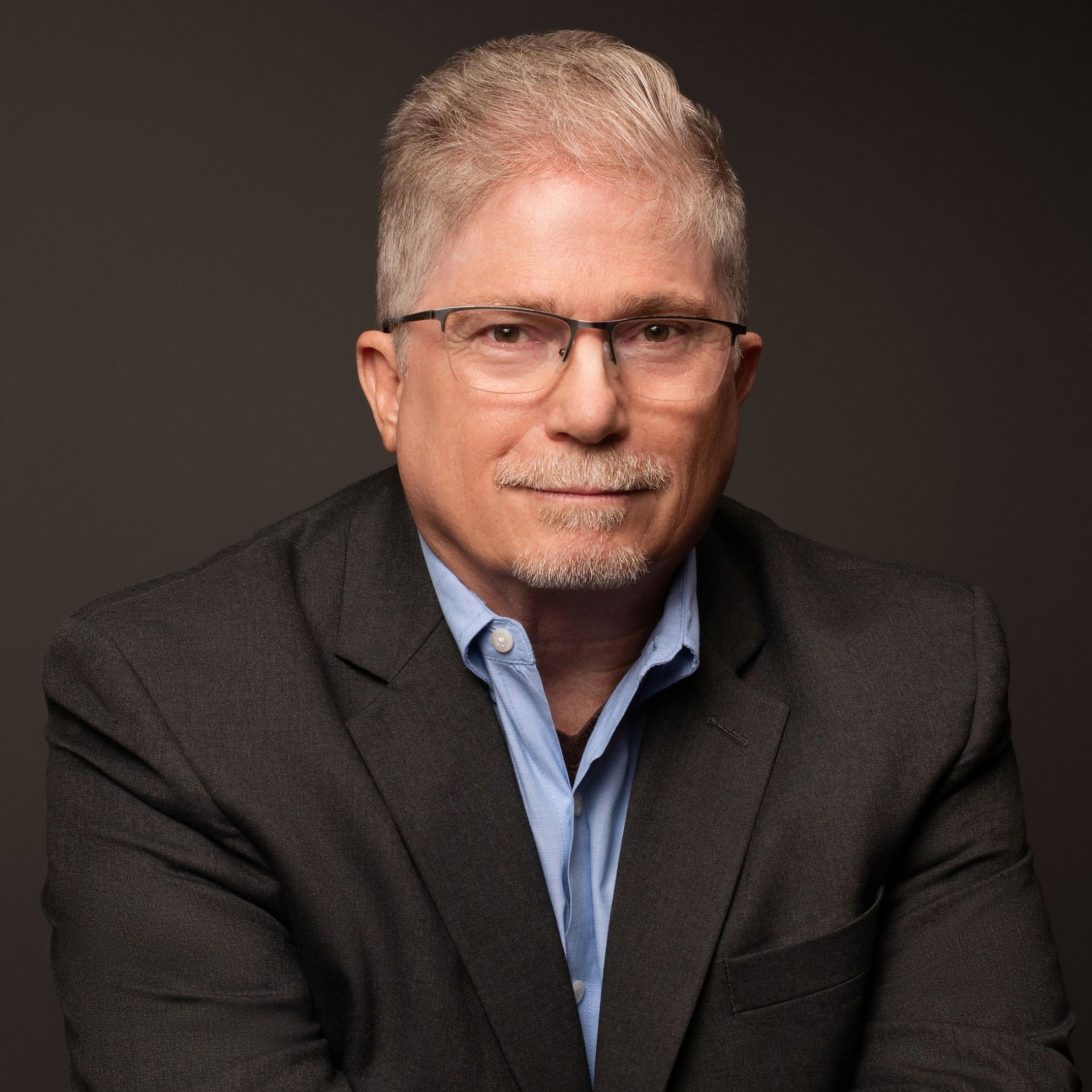 Man with glasses and grey hair wearing a blazer and open-collared blue shirt against a grey background.