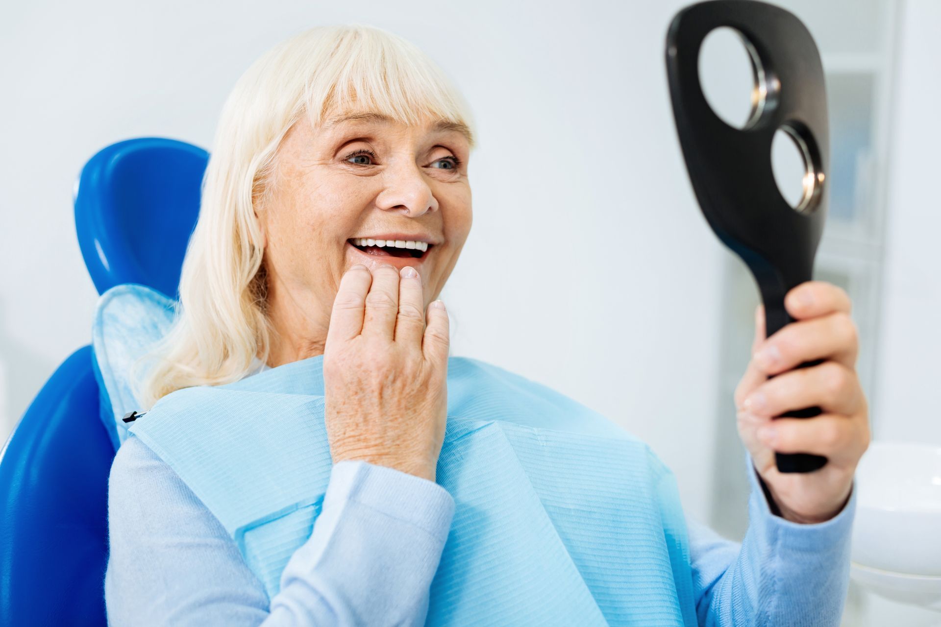 an elderly woman in a dental chair looking at her teeth in a mirror.