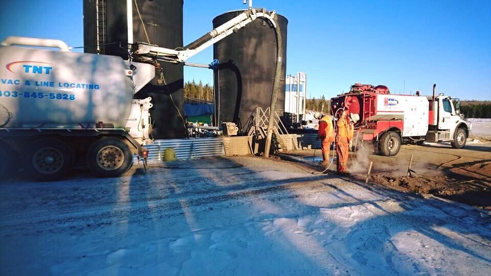 Workers in high-visibility gear stand near a vacuum truck and large storage tanks at a snowy industrial work site.