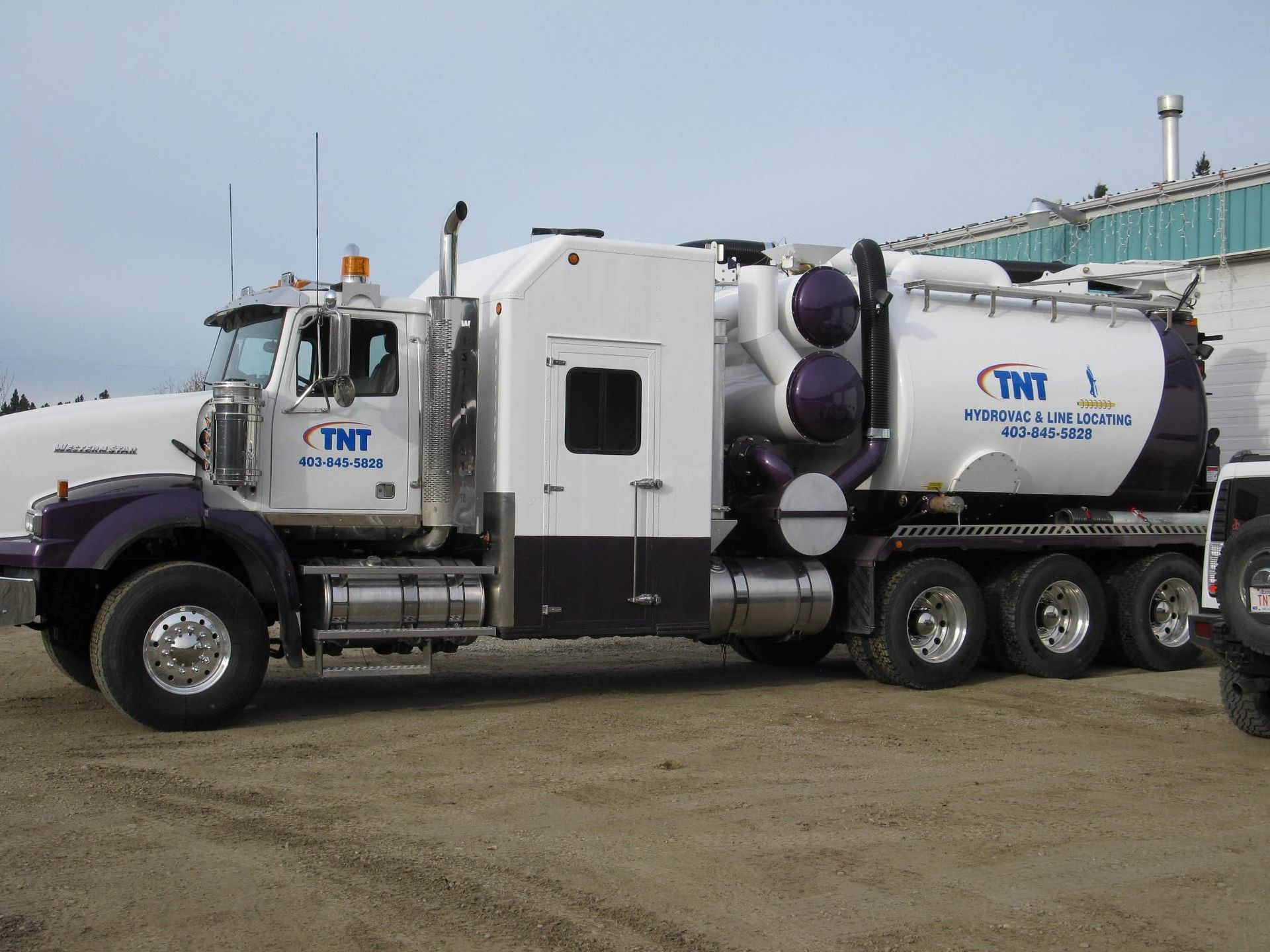 A white and purple vacuum truck with TNT branding parked on a gravel lot under an overcast sky.