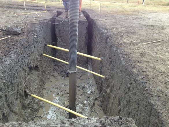 A deep trench in muddy ground features wooden shoring braces spanning the walls and a central vertical support pipe.