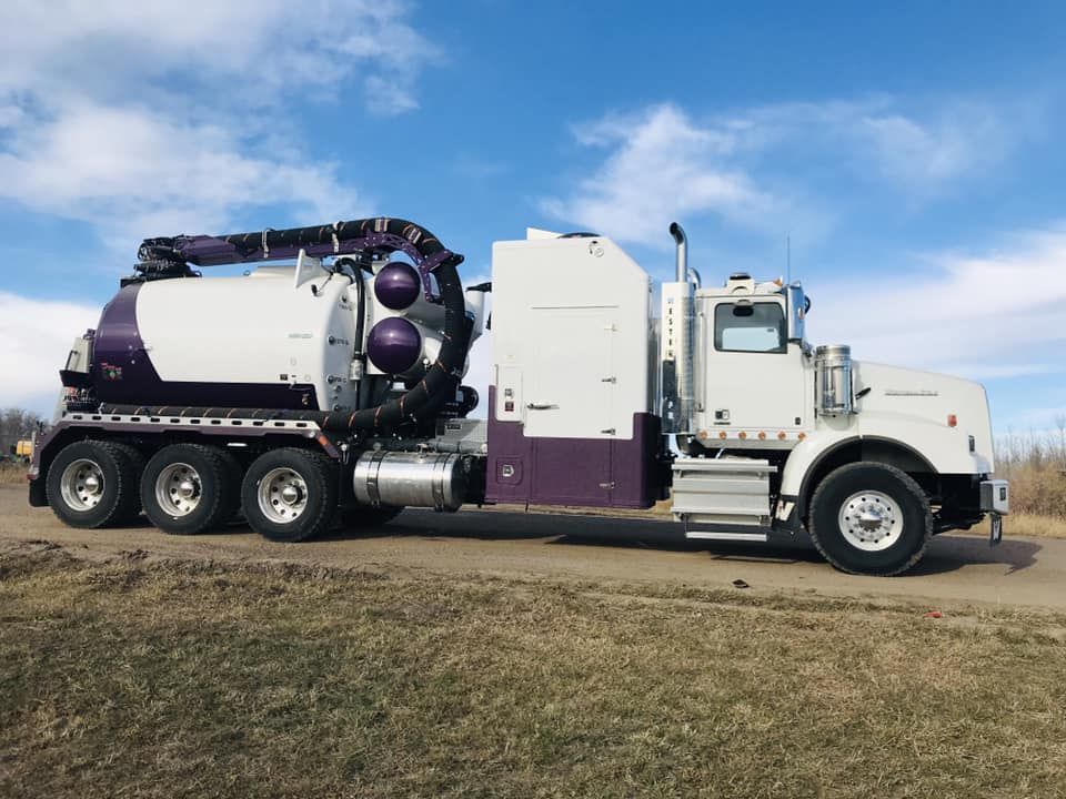 A white and purple vacuum excavation truck parked on a dirt road against a blue sky with clouds.