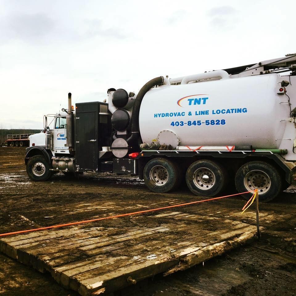 A white TNT Hydrovac & Line Locating truck parked on a muddy work site next to wooden mats.