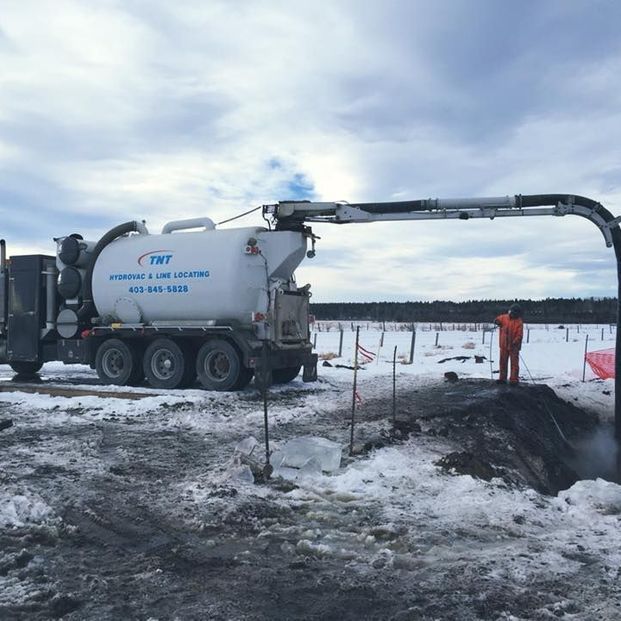 A vacuum excavation truck on a snowy construction site uses a boom to clear a trench as a worker stands nearby.