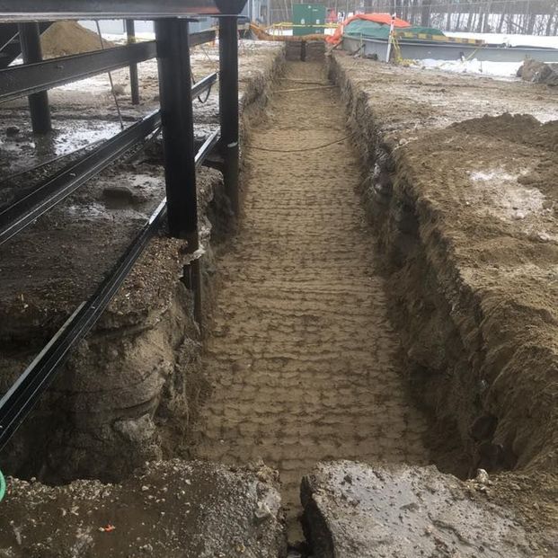 A view looking down a narrow trench excavated in dirt next to a metal structural support frame at a construction site.
