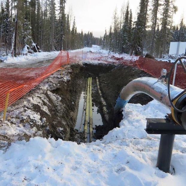 A construction trench with exposed yellow pipes, surrounded by snow, orange fencing, and a forested backdrop.