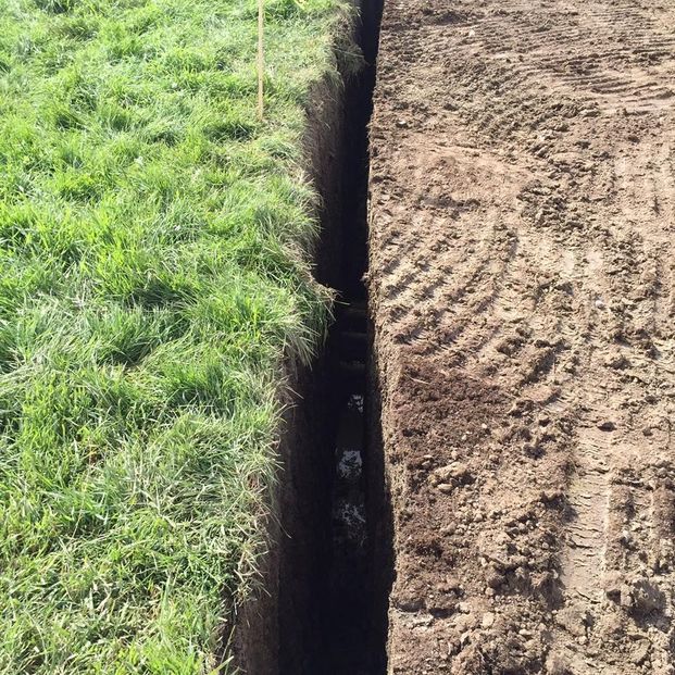 A deep, narrow trench dug into the ground, separating a grassy field on the left from tilled, exposed earth on the right.