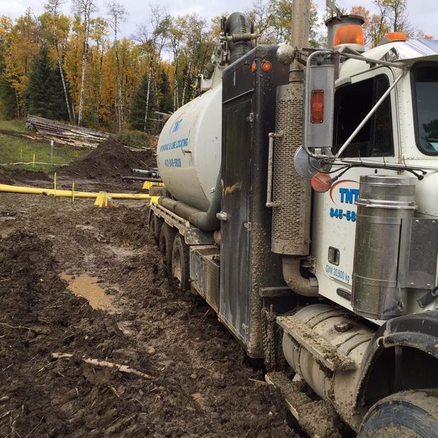 A large white industrial vacuum truck parked on muddy ground at an outdoor work site with trees in the background.