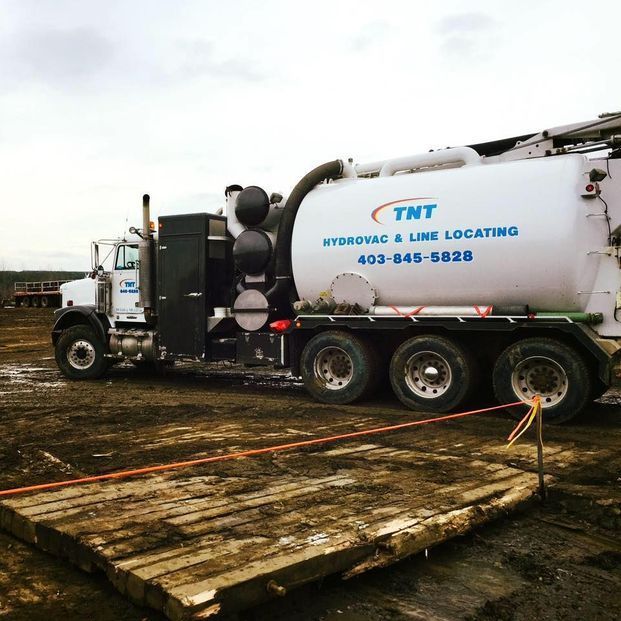A white TNT hydrovac truck parked on a muddy construction site, positioned on a wooden platform.