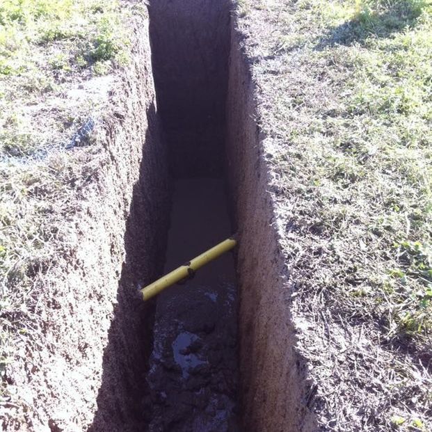 A deep, narrow dirt trench in a grassy area with a single yellow utility pipe running across the bottom.