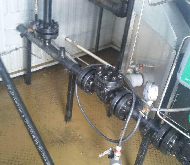 Industrial plumbing system with black metal pipes, flanged valves, and pressure gauges in an indoor utility room.