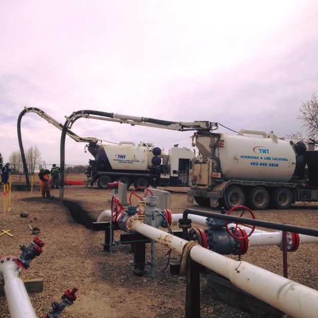 Two vacuum excavation trucks work at a pipeline site with industrial pipes and valves in the foreground.