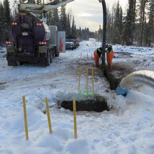 Workers use a vacuum truck in a snowy landscape to excavate soil near utility markers and a large pipe.