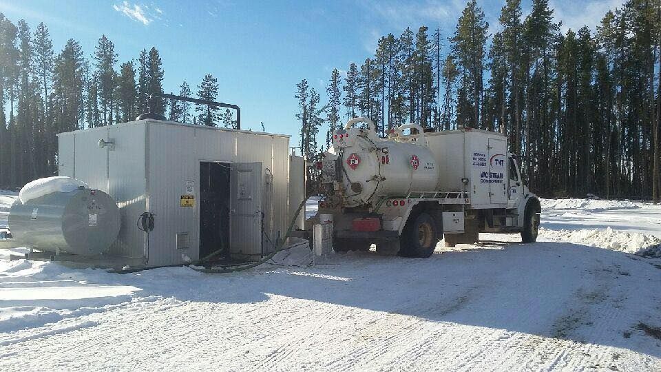 A white tanker truck parked in the snow next to a metal equipment enclosure in a forest clearing.