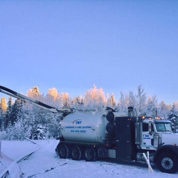A white vacuum truck with a long extendable boom parked in a snowy, sunlit forest clearing.