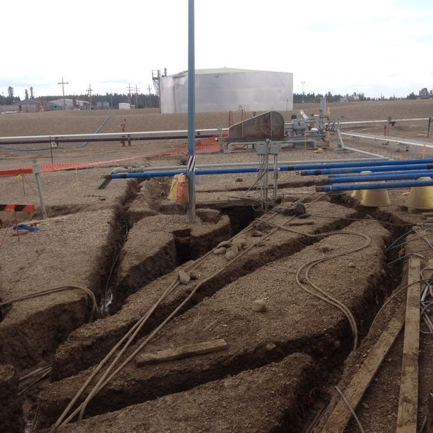 A construction site features open trenches dug into dry, dusty ground near blue pipes and a distant storage tank.