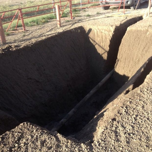 A deep rectangular trench excavated in dirt, featuring a utility pipe running across the bottom of the pit.