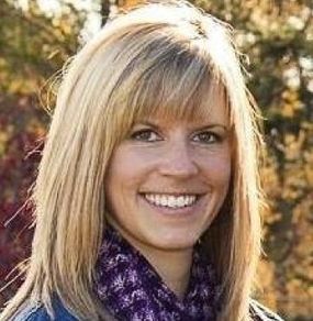 A smiling blonde person wearing a patterned purple scarf with an out-of-focus outdoor background.