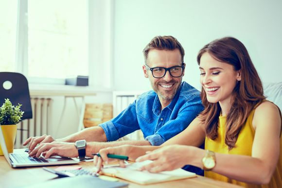 Two coworkers smiling and working together at a desk with papers, laptop, and plant in a bright office