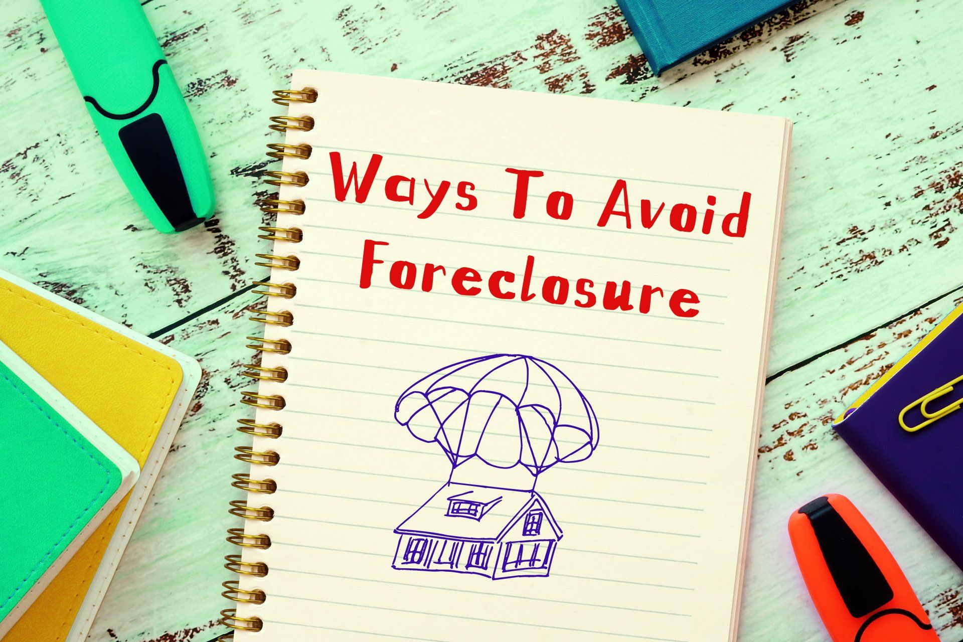 Notebook page reading “Ways To Avoid Foreclosure” with a small house sketch on a desk surrounded by colorful stationery