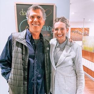 Two people smiling indoors in front of a chalkboard and hallway wall art