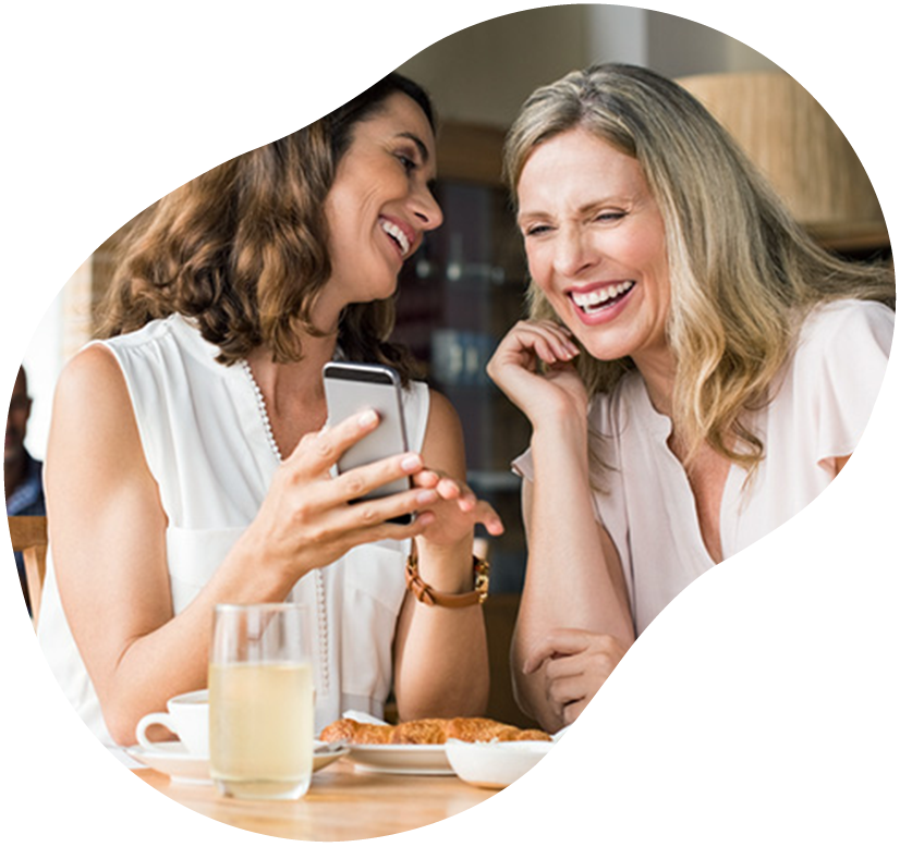 Two women smiling at a café while looking at a smartphone beside drinks and pastries
