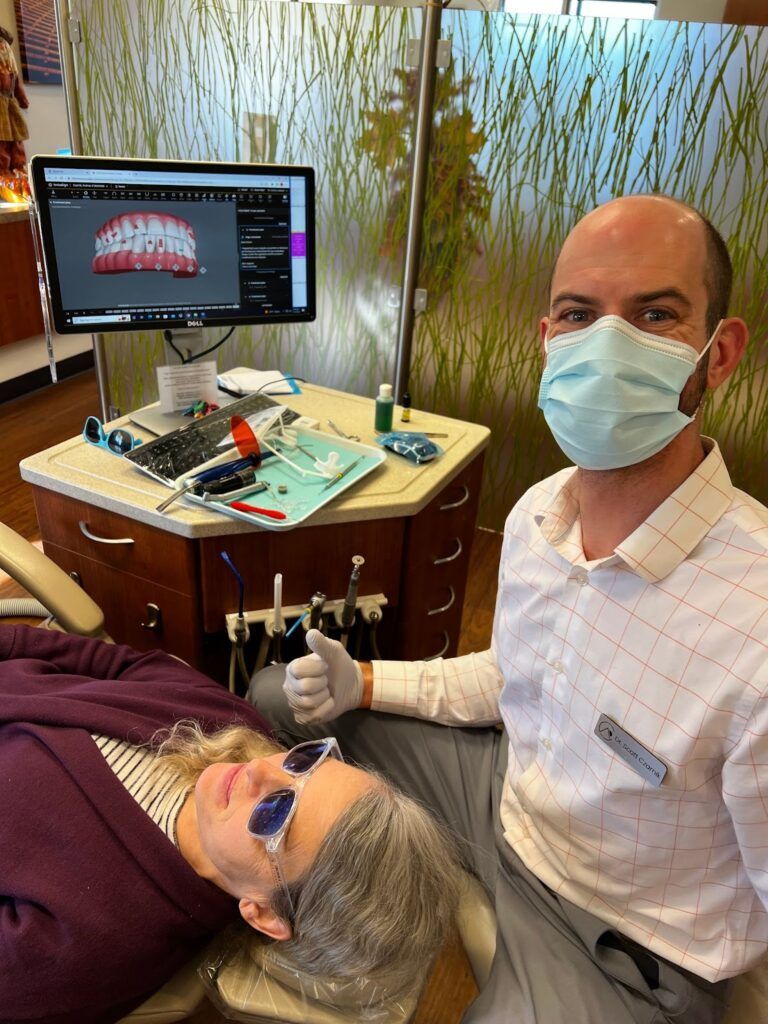 Dentist wearing a mask works on a patient reclined in a chair beside a monitor showing dental X-rays.