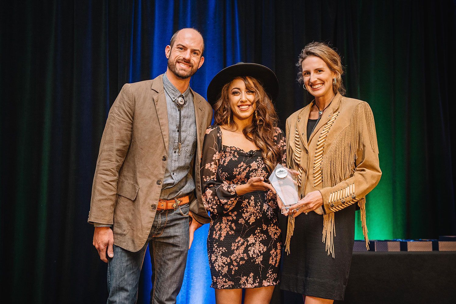 Three people posing on a stage, holding an award, with dark curtains and green lighting behind them.
