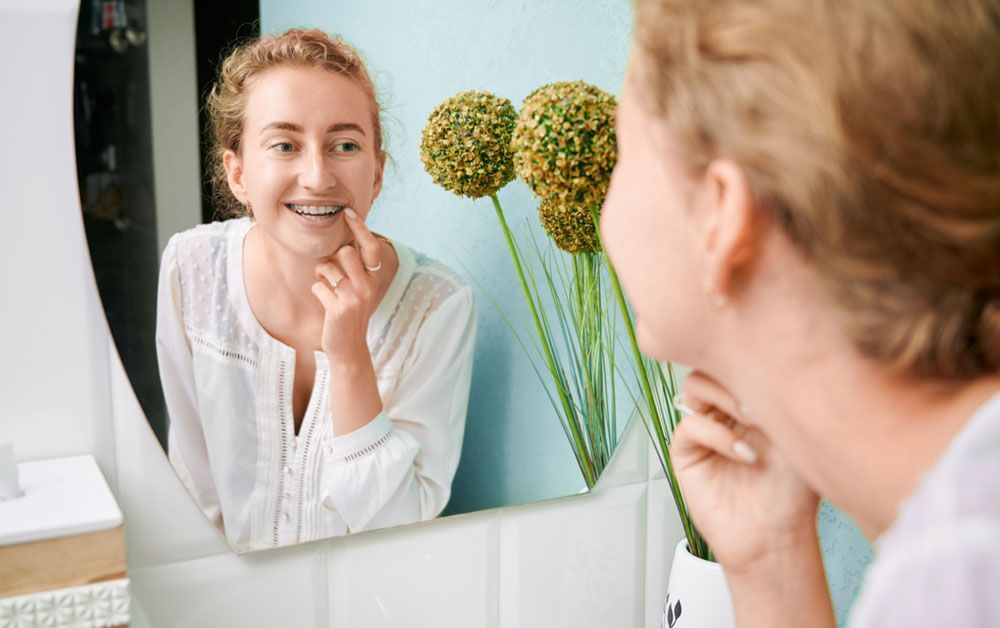 Person smiling at bathroom mirror, touching their chin while inspecting skin; white robe and green flowers in vase.