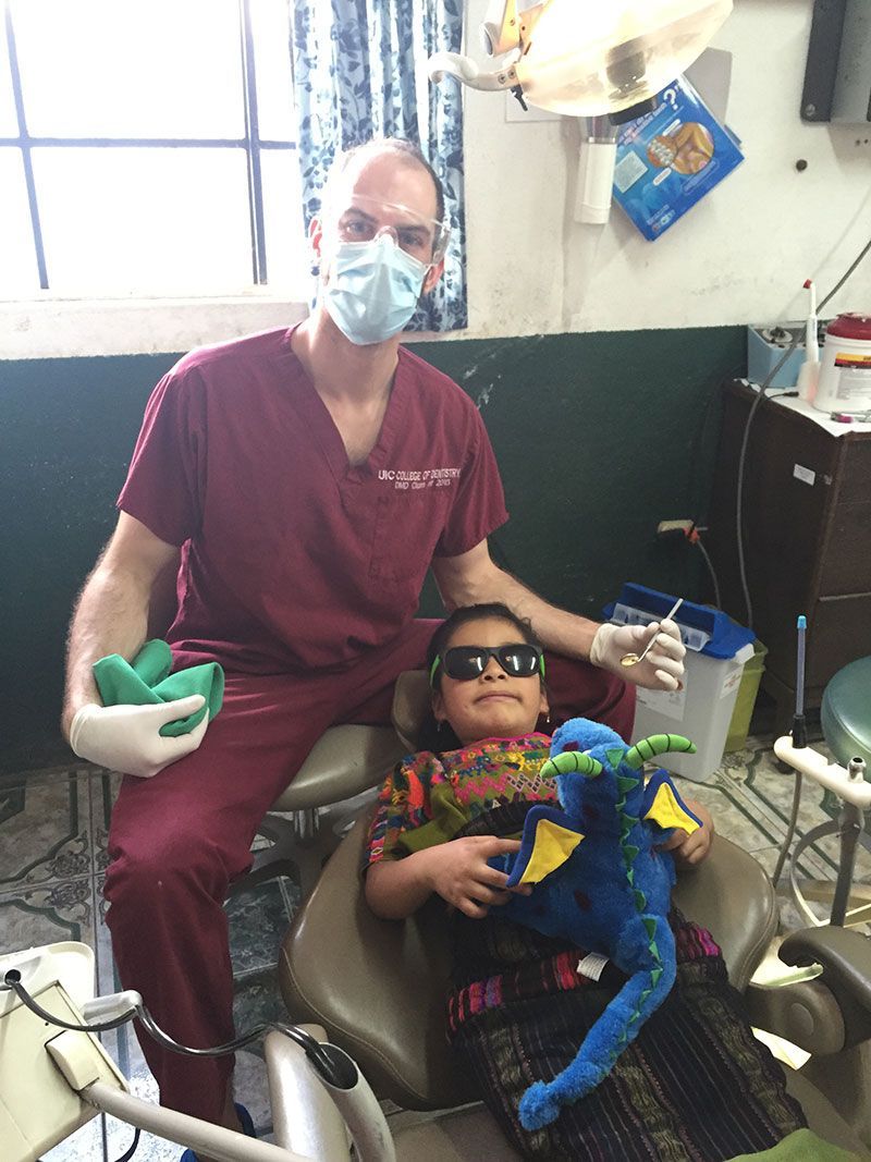 Dentist and child in exam chair, both wearing masks; child holds a blue plush toy.