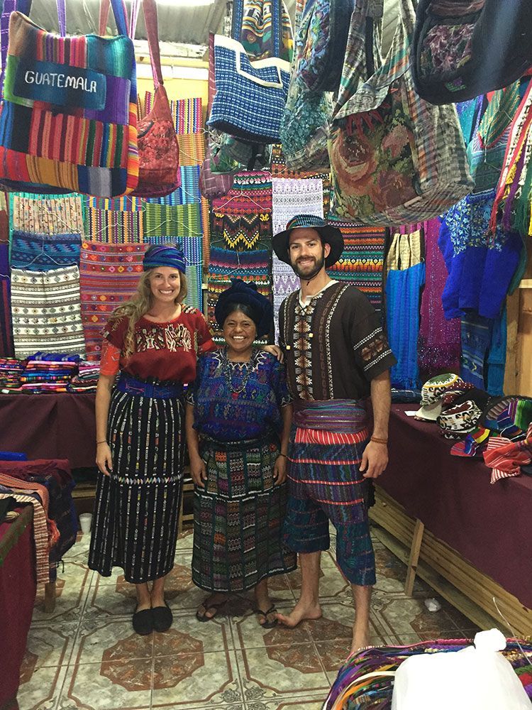 Three people standing in a colorful textile market stall, surrounded by hanging woven bags and rugs