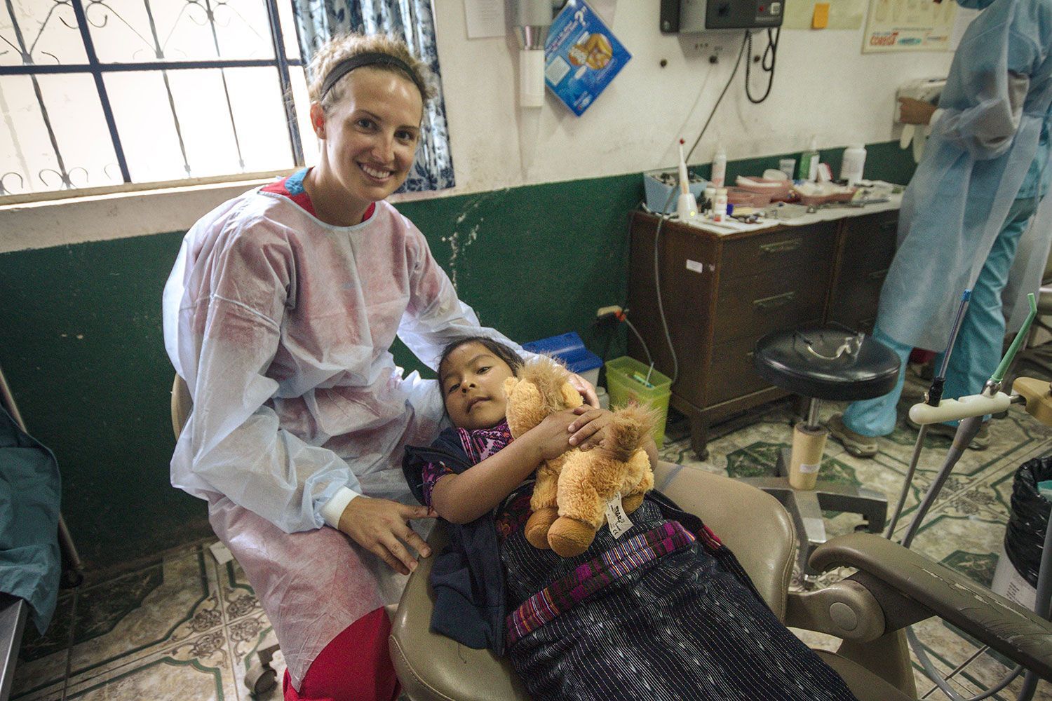 A woman in a pink gown sits beside a child lying on a hospital bed, holding a teddy bear.