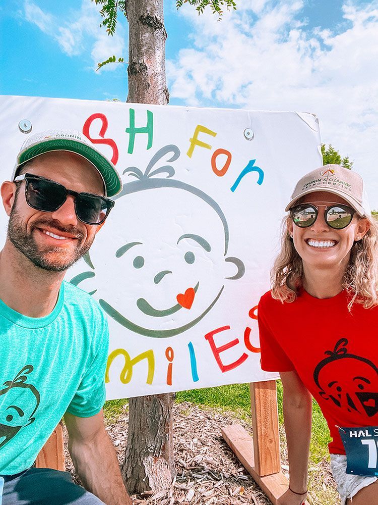 Two people smiling beside a colorful “Shoe to Smile” sign outdoors