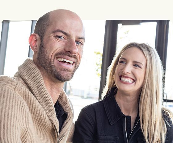 Two smiling people seated indoors, with a bald man in a beige sweater and a blonde woman in a black shirt
