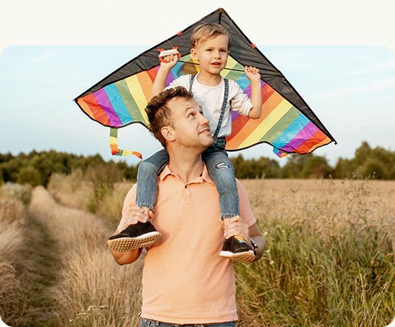 Child on a man’s shoulders flying a colorful kite in a grassy field