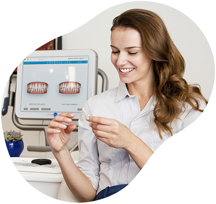 Woman smiling while holding a dental aligner beside a computer showing teeth images