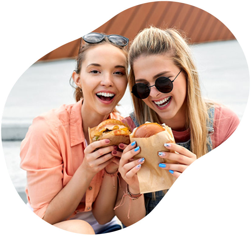 Two smiling friends eating burgers outdoors, holding food close and laughing in a casual setting