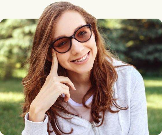 Smiling person in sunglasses with a hand on their cheek, outdoors with blurred greenery behind.