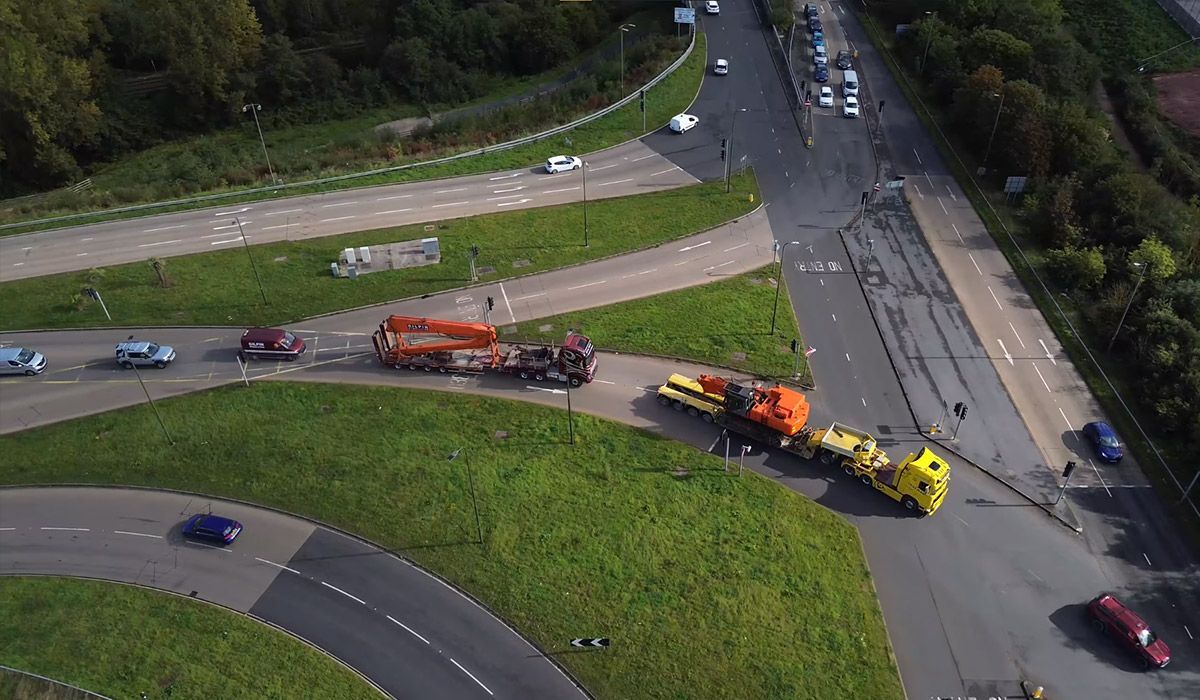 Aerial view of two large trucks navigating a roundabout, with other cars and greenery visible.