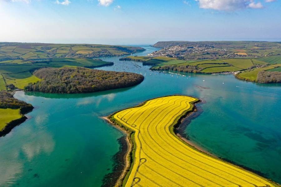 Aerial view of a bright blue river with yellow fields and green hills leading to a town by the sea on a sunny day.