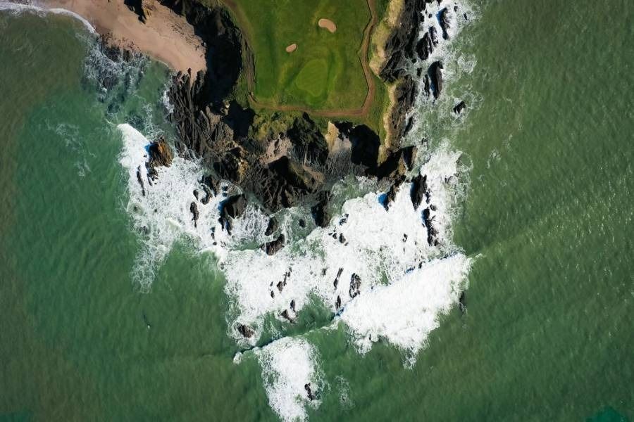 Overhead view of a rocky coastline with foamy waves and a green golf course.