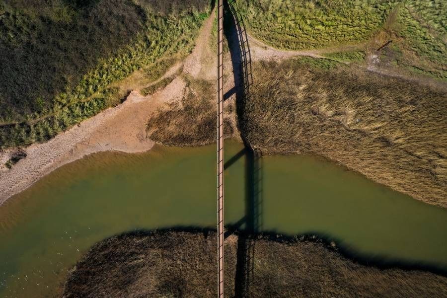 Overhead view of a bridge crossing a river, with marshland on either side, casting a shadow.