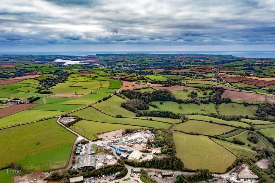 Aerial view of green fields, trees, and buildings under a cloudy sky, near a coastline.