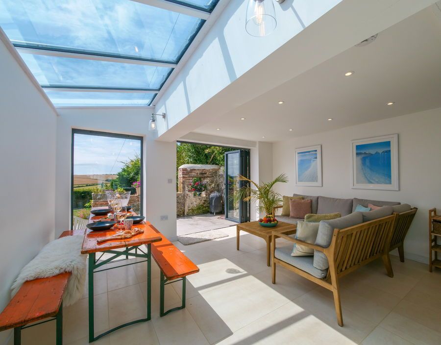 Bright sunroom with glass roof and open doors.  Dining table, wicker furniture, and artwork.