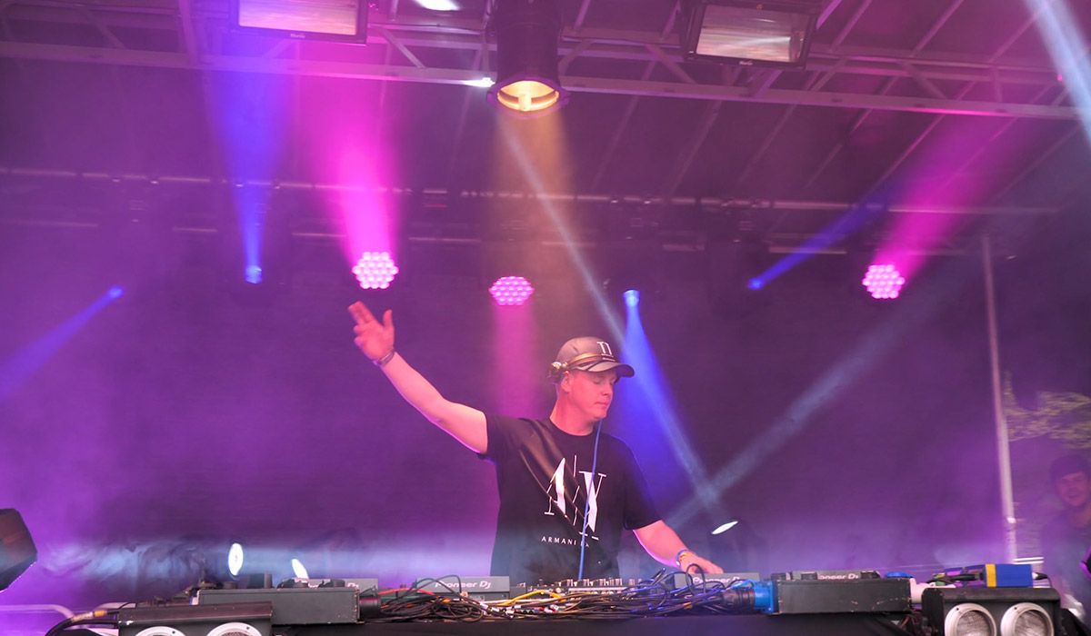 DJ on stage, waving, operating equipment under purple and pink stage lights and smoke.