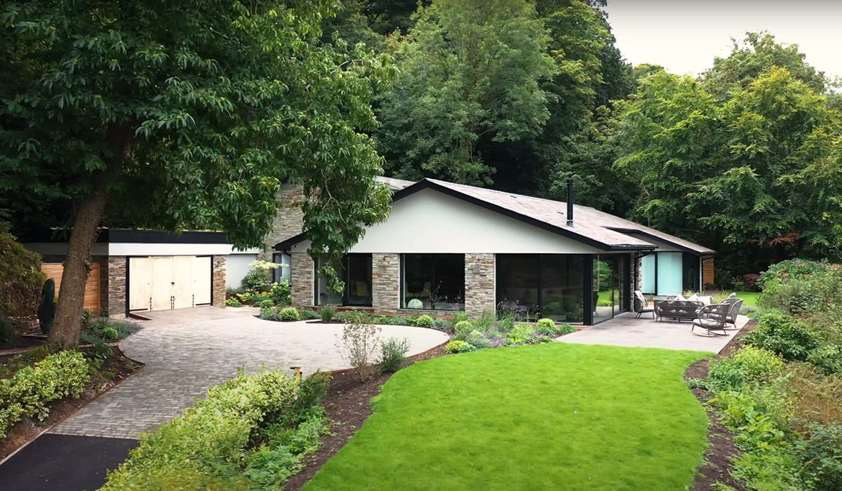 Mid-century modern house nestled amongst trees with a stone facade, flat roof, and a paved driveway.