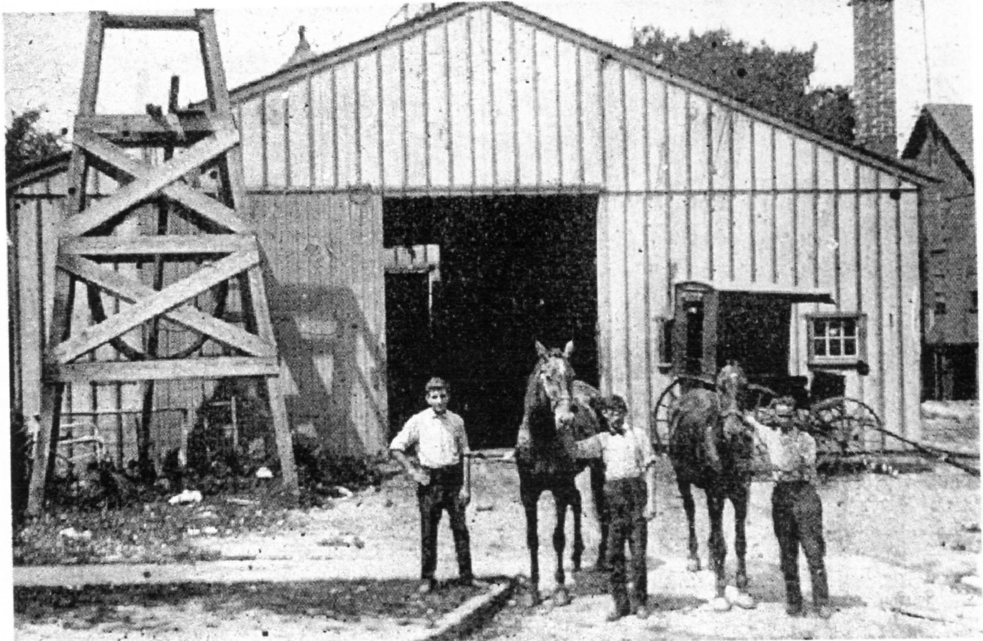Young men with horses outside a wooden barn, from the horse-drawn era.